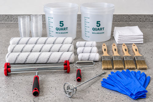 A paint roller kit arranged on a gray countertop, including two large white 5-quart buckets, clear cups, white cloths, white roller sleeves, four paint brushes with tan bristles, a metal mixing paddle, a roller attachment, and a pair of blue gloves.